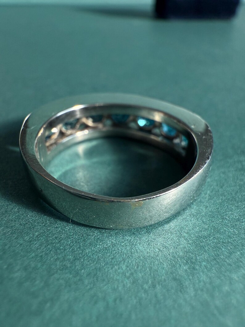 May include: A silver ring with a row of small, bright blue gemstones set into the band. The ring is shown on a teal-colored surface, with the focus on the gemstones and the polished metal.