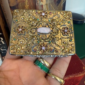 Può includere: Scatola rettangolare dorata con intricati disegni floreali e a volute. La scatola presenta una pietra centrale ovale e opalescente ed è impreziosita da gemme colorate. I bordi sono argentati. Una mano con diversi anelli tiene la scatola.