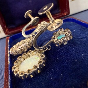 May include: Close-up of vintage silver screw-back earrings. Each earring features an oval, iridescent opal center stone surrounded by a decorative silver setting. One earring has a small, round, blue-green stone. The earrings are displayed in a blue velvet-lined box.