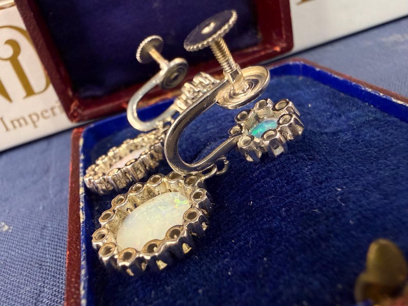 May include: Close-up of vintage silver screw-back earrings. Each earring features an oval, iridescent opal center stone surrounded by a decorative silver setting. One earring has a small, round, blue-green stone. The earrings are displayed in a blue velvet-lined box.