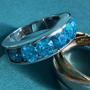 May include: A silver ring with five oval-cut, bright blue gemstones. The ring is set against a teal background, with a gold ring partially visible in the foreground. The gemstones are a vibrant contrast to the silver band.