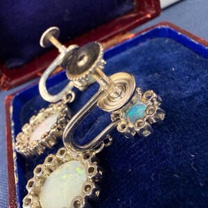 May include: Close-up of vintage silver-tone earrings with opalescent stones. The earrings have a screw-back design and are set in a decorative, floral-inspired frame. The opals display a play of color, with hints of blue and green.