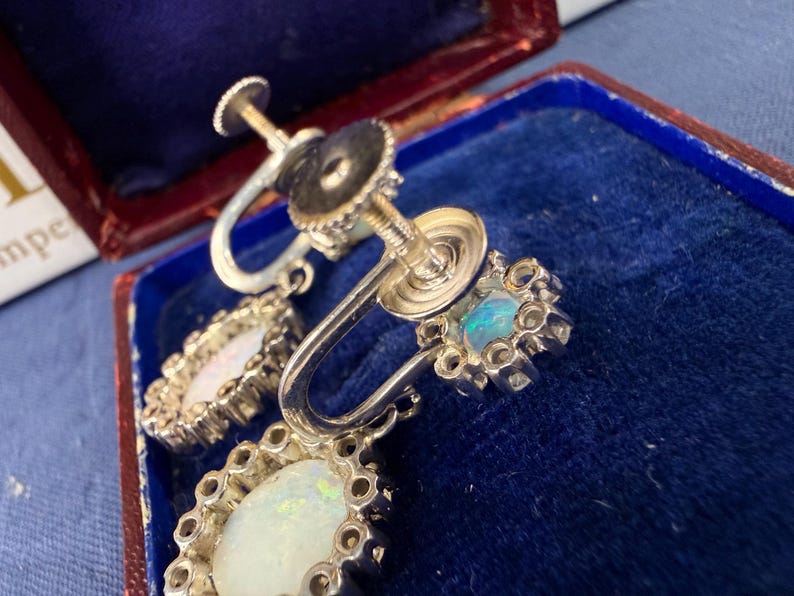 May include: Close-up of vintage silver-tone earrings with opalescent stones. The earrings have a screw-back design and are set in a decorative, floral-inspired frame. The opals display a play of color, with hints of blue and green.