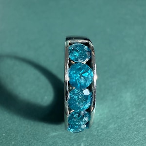 May include: A silver ring featuring five round, faceted, bright blue gemstones. The gemstones are set closely together, creating a continuous band of color. The ring is displayed against a teal background.
