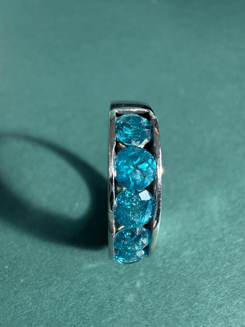 May include: A silver ring featuring five round, faceted, bright blue gemstones. The gemstones are set closely together, creating a continuous band of color. The ring is displayed against a teal background.