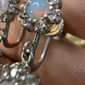 May include: Close-up of a pair of silver earrings. Each earring features a central, iridescent opal stone surrounded by clear, sparkling accent stones. The earrings have a floral design.