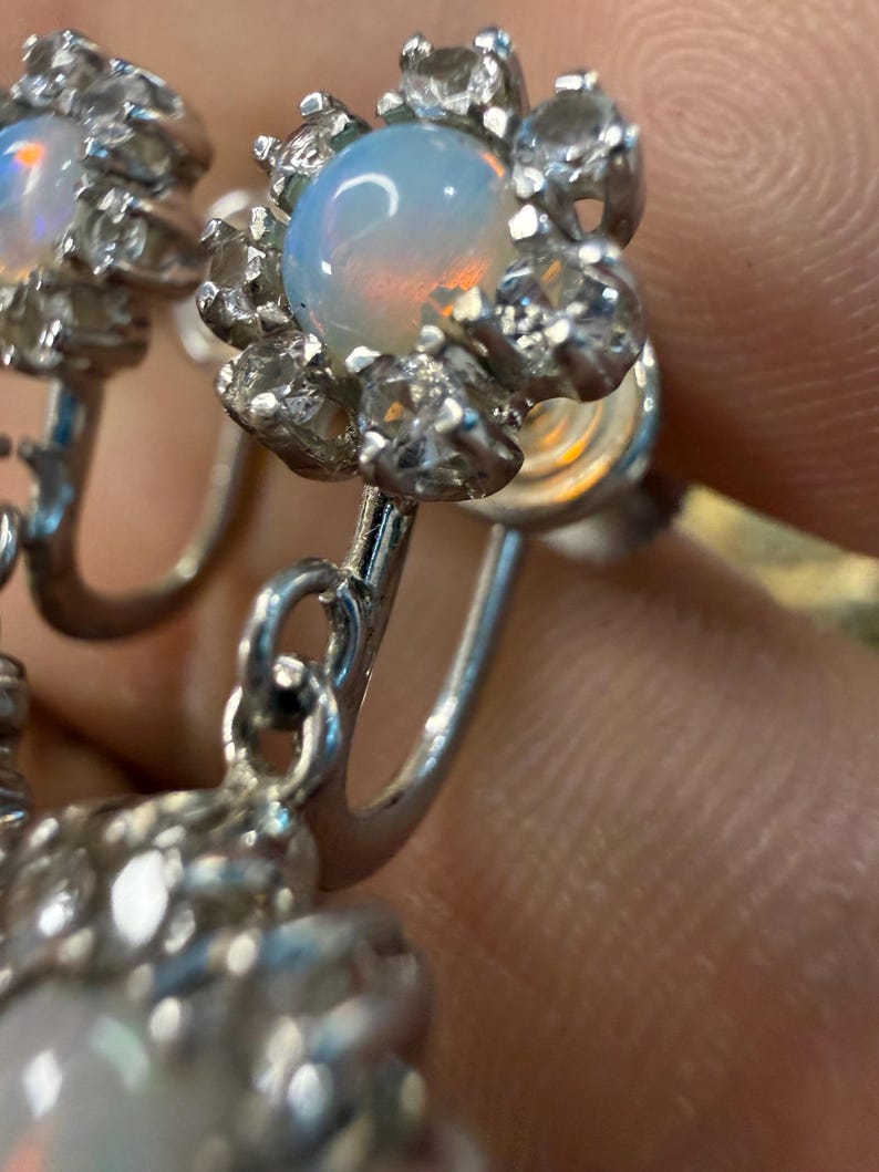 May include: Close-up of a pair of silver earrings. Each earring features a central, iridescent opal stone surrounded by clear, sparkling accent stones. The earrings have a floral design.