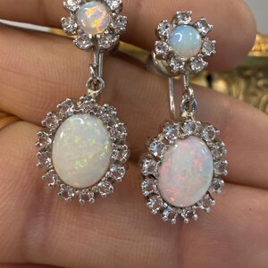 May include: A pair of silver-toned earrings featuring an oval, iridescent opal stone surrounded by a halo of small, clear crystals. The earrings have a floral design at the top and a clip-on style.