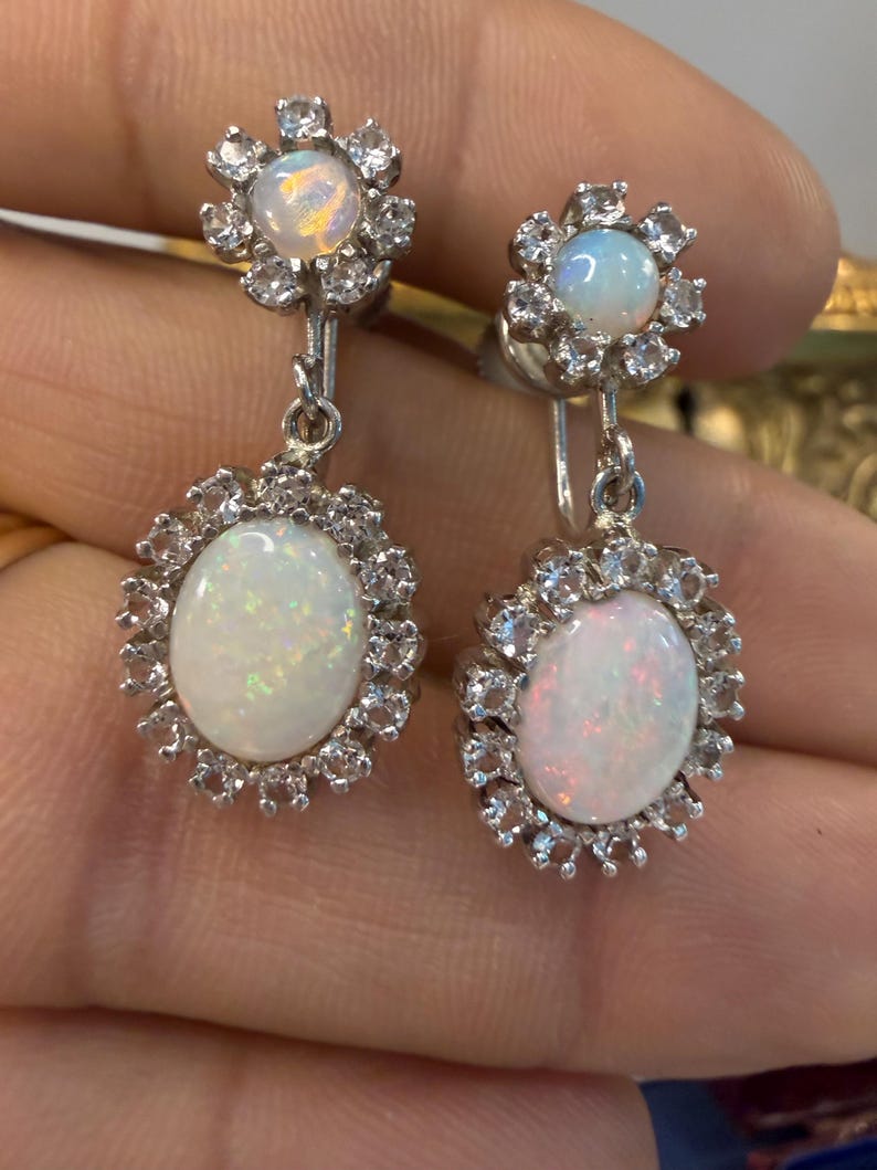 May include: A pair of silver-toned earrings featuring an oval, iridescent opal stone surrounded by a halo of small, clear crystals. The earrings have a floral design at the top and a clip-on style.