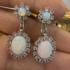 May include: A pair of silver drop earrings featuring oval opal stones surrounded by a halo of small, clear gemstones. The earrings have a floral design at the top and are held by a silver hook.