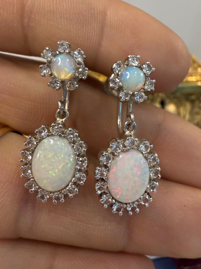 May include: A pair of silver drop earrings featuring oval opal stones surrounded by a halo of small, clear gemstones. The earrings have a floral design at the top and are held by a silver hook.