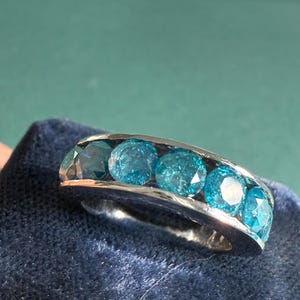 May include: A silver ring featuring five round, bright blue gemstones. The gemstones are set in a row, creating a continuous band of color. The ring is displayed on a dark blue, textured surface.