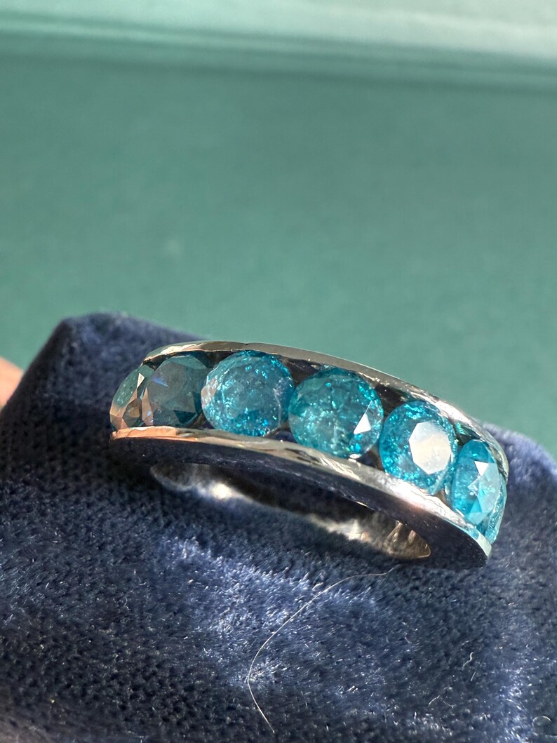 May include: A silver ring featuring five round, bright blue gemstones. The gemstones are set in a row, creating a continuous band of color. The ring is displayed on a dark blue, textured surface.