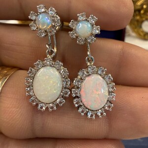 May include: A pair of silver drop earrings featuring opalescent oval-shaped stones surrounded by small, clear gemstones. The earrings have a floral design at the top and a larger oval stone at the bottom, creating a classic, elegant look.