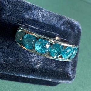 May include: A silver ring featuring a row of six round, bright teal gemstones. The ring is set against a dark blue velvet surface, with a hint of a green background. The gemstones are evenly spaced and catch the light.