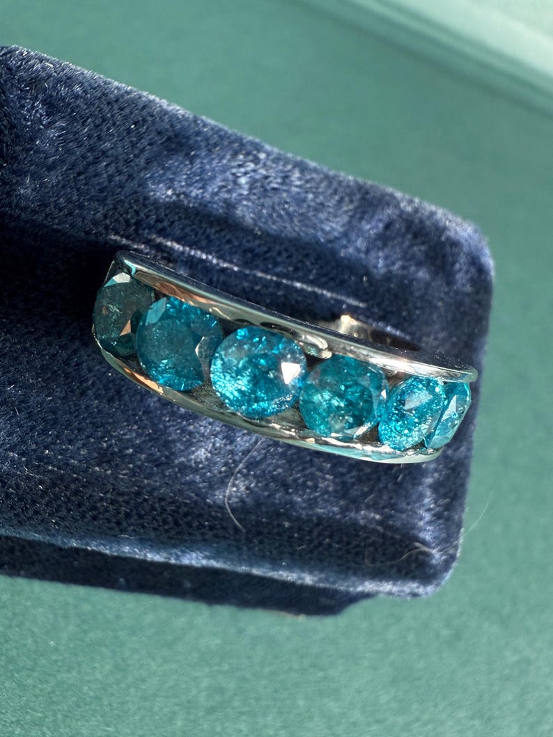 May include: A silver ring featuring a row of six round, bright teal gemstones. The ring is set against a dark blue velvet surface, with a hint of a green background. The gemstones are evenly spaced and catch the light.