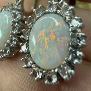 May include: Close-up of a pair of silver earrings. Each earring features a large, oval opal stone surrounded by a halo of small, clear gemstones. The opals display iridescent colors, and the silver setting has intricate details.