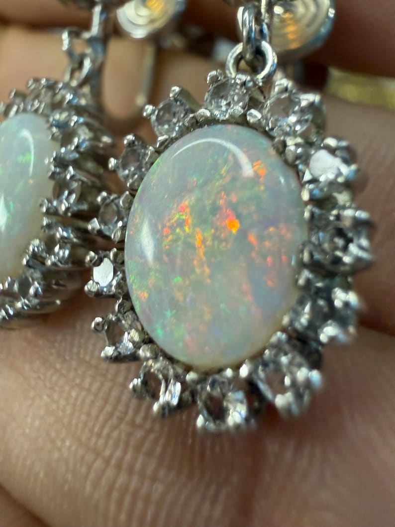May include: Close-up of a pair of silver earrings. Each earring features a large, oval opal stone surrounded by a halo of small, clear gemstones. The opals display iridescent colors, and the silver setting has intricate details.