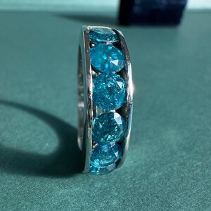 May include: A silver ring featuring a row of round, faceted, bright blue gemstones. The ring is set against a teal-green background, with a dark blue velvet ring box in the background.