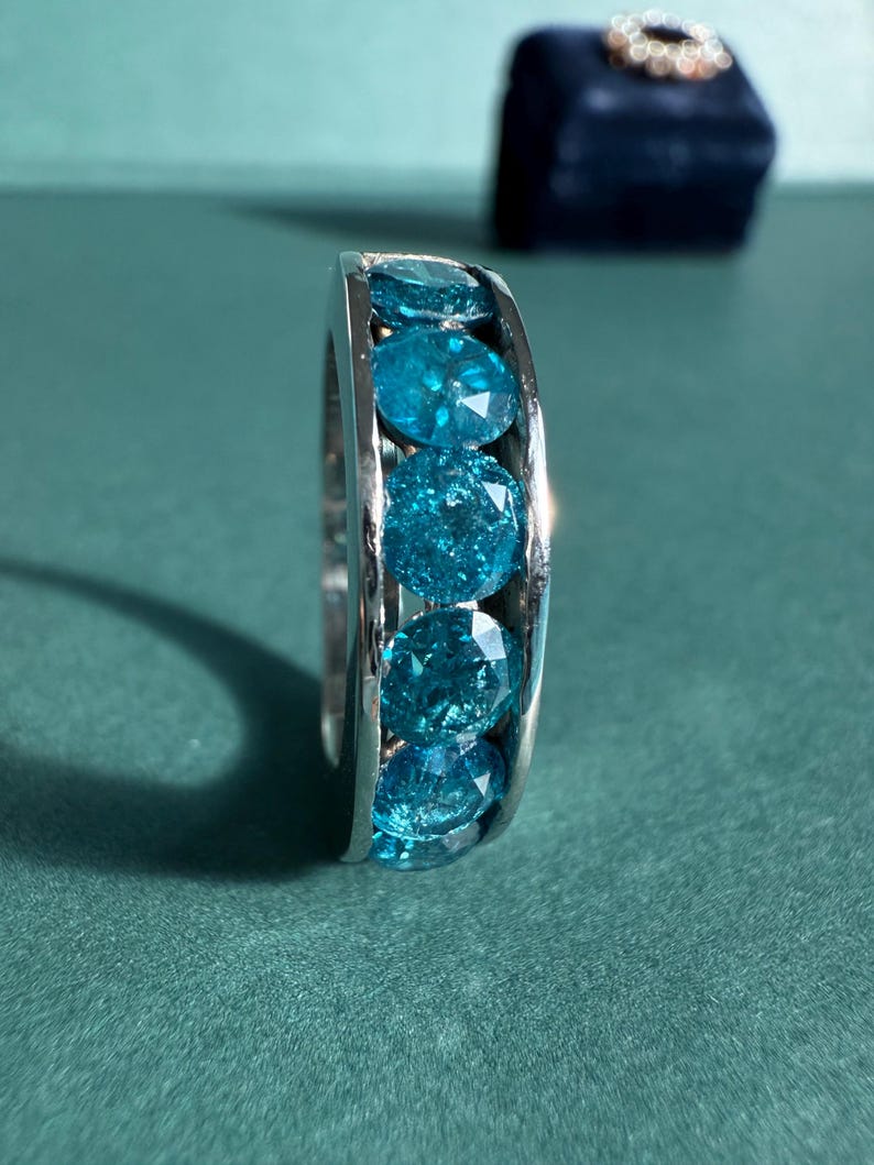 May include: A silver ring featuring a row of round, faceted, bright blue gemstones. The ring is set against a teal-green background, with a dark blue velvet ring box in the background.