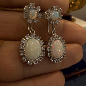 May include: A pair of silver-toned earrings featuring opalescent oval-shaped stones surrounded by a halo of smaller, clear stones. The earrings have a floral design at the top and a dangling oval opal at the bottom.