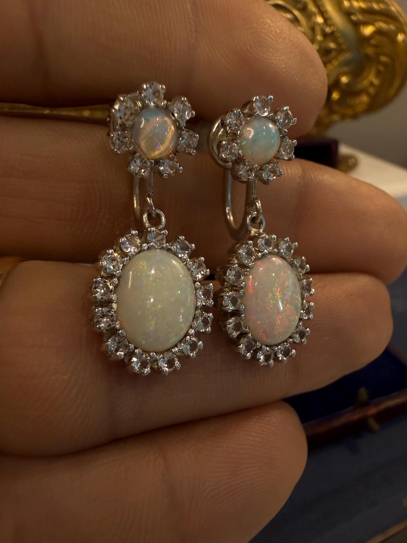 May include: A pair of silver-toned earrings featuring opalescent oval-shaped stones surrounded by a halo of smaller, clear stones. The earrings have a floral design at the top and a dangling oval opal at the bottom.
