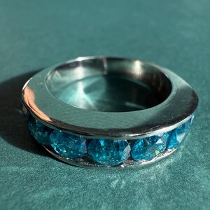 May include: A silver-toned ring with a band of round, bright blue gemstones. The ring is set against a teal background, with the gemstones catching the light. The ring is a piece of jewelry.