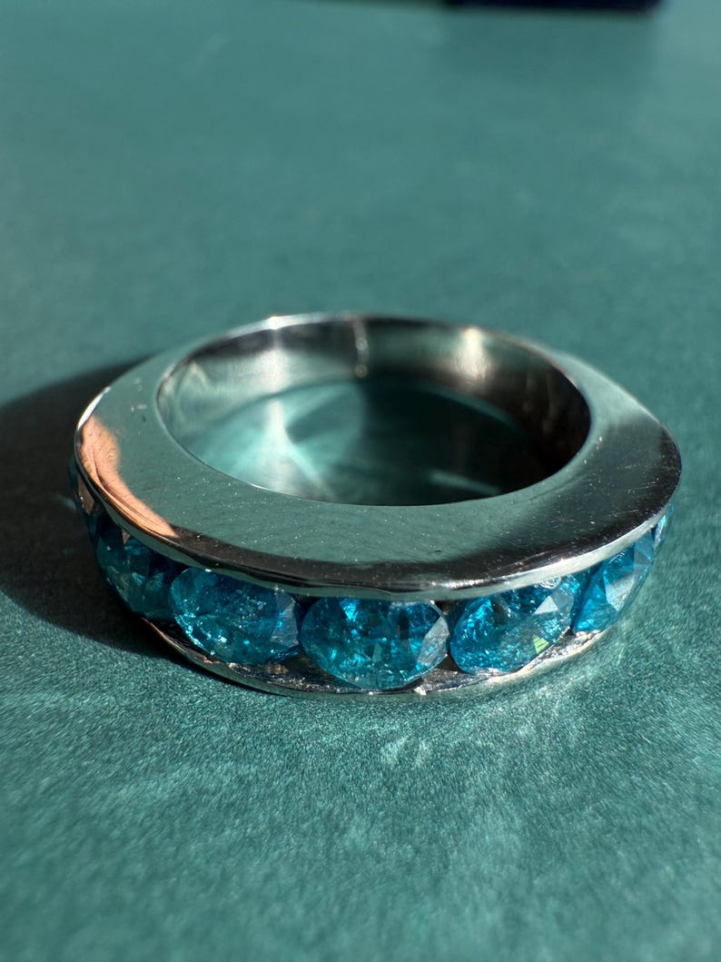 May include: A silver-toned ring with a band of round, bright blue gemstones. The ring is set against a teal background, with the gemstones catching the light. The ring is a piece of jewelry.