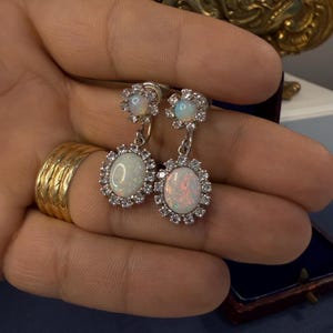 May include: Silver-toned drop earrings featuring oval opal stones surrounded by a halo of small, clear crystals. The earrings have a floral design at the top and are held in a hand.