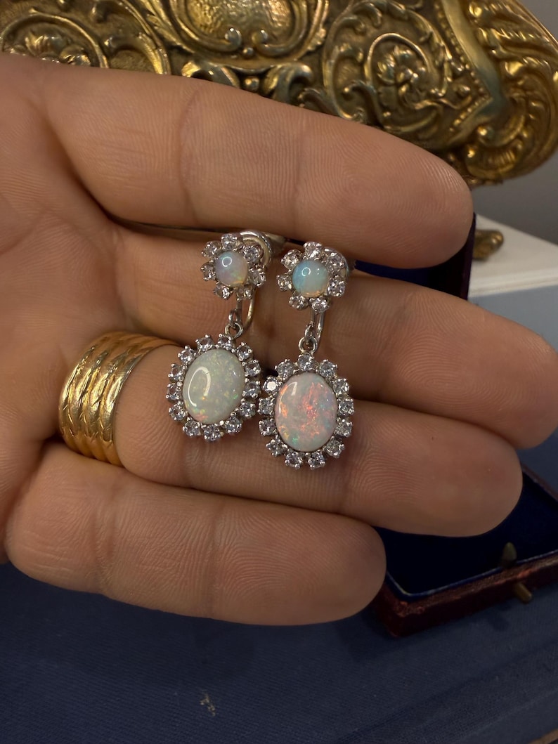 May include: Silver-toned drop earrings featuring oval opal stones surrounded by a halo of small, clear crystals. The earrings have a floral design at the top and are held in a hand.