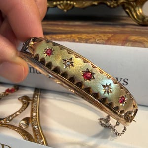 May include: A gold hinged bangle bracelet featuring a scalloped edge design. The surface is adorned with alternating star-set red gemstones and clear stones. The piece includes a delicate safety chain attached to the side of the metal frame.