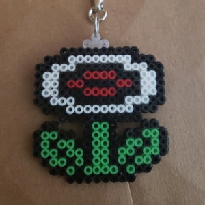 Könnte beinhalten: Ein schwarzer, weißer, roter und grüner pixelartiger Blumen-Schlüsselanhänger. Die Blume ist eine weiße und schwarze Blume mit einer roten Mitte. Die Blume hat einen grünen Stiel und Blätter.