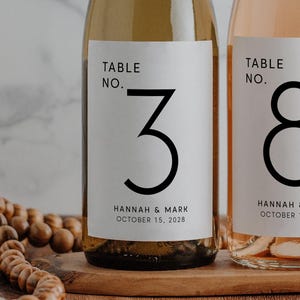 May include: Two wine bottles with labels reading "Table No. 3" and "Table No. 8" with the names "Hannah & Mark" and the date "October 15, 2028". The bottles are on a wooden tray, with a marble background.