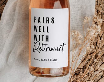 May include: A bottle of rosé wine with a gold cap and a white label that reads "Pairs Well With Retirement." The bottle is set on a woven placemat with a white cloth and dried flowers.