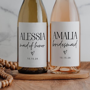May include: Two wine bottles with custom labels. One bottle is filled with white wine, the other with rosé. The labels read "Alessia maid of honor" and "Amalia bridesmaid" with a date. The bottles sit on a wooden tray.