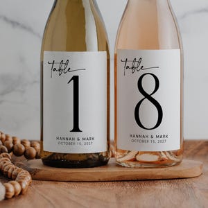 May include: Two wine bottles with labels for a wedding. One bottle is filled with white wine and the other with rosé. The labels read "Table 1" and "Table 8" with the names "Hannah & Mark" and the date "October 15, 2027".