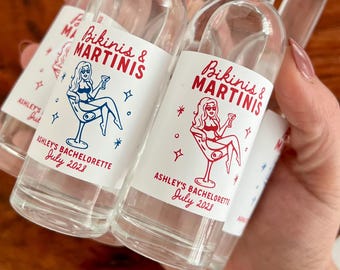 May include: Small clear glass bottles with white screw caps, each featuring a white label printed with red and blue text reading Bikinis & Martinis, Ashley's Bachelorette, July 2023, and a graphic of a person sitting in a martini glass.
