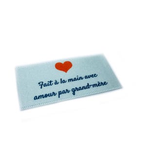Peut inclure: Une étiquette en tissu bleu clair avec un cœur rouge et le texte "Fait à la main avec amour par grand-mère".
