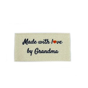 Peut inclure: Une étiquette en tissu beige avec le texte "Made with love by Grandma" en écriture cursive noire. Un petit cœur rouge est inclus dans le texte.