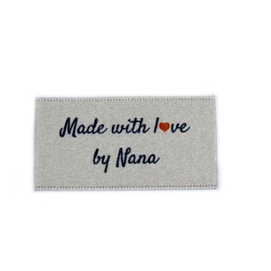 Könnte beinhalten: Ein beigefarbenes Stofflabel mit dem Text "Made with love by Nana" in dunkelblauer Schrift und einem kleinen roten Herz.