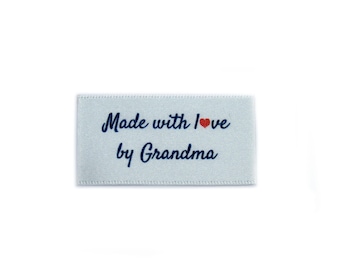 Puede incluir: Etiqueta de satén blanco con texto azul que dice "Made with love by Grandma" con un pequeño corazón rojo.