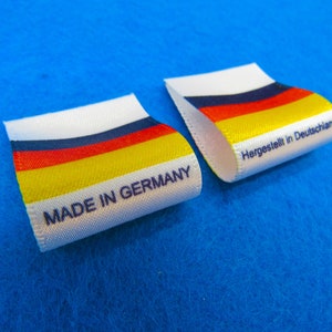 May include: Two white fabric labels with black text. One label reads "Made in Germany" and the other reads "Hergestellt in Deutschland". Both labels feature a horizontal stripe pattern in the colors of the German flag: black, red, yellow, and white.