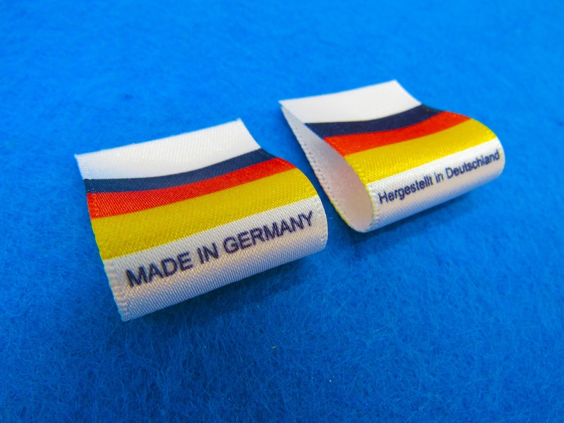 May include: Two white fabric labels with black text. One label reads "Made in Germany" and the other reads "Hergestellt in Deutschland". Both labels feature a horizontal stripe pattern in the colors of the German flag: black, red, yellow, and white.