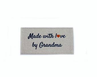 Puede incluir: Una etiqueta de tela beige con el texto "Made with love by Grandma" en letra cursiva azul y un pequeño corazón rojo.