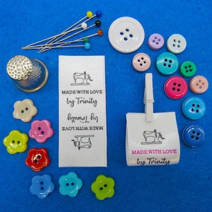 Op de afbeelding: Twee witte satijnen labels met zwarte naaimachine-afbeeldingen en de tekst "MADE WITH LOVE by Trinity" in zwart en roze. De labels zijn omringd door kleurrijke knopen en naalden.