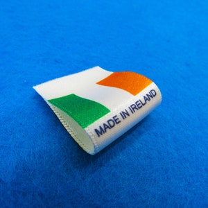 Könnte beinhalten: Weißes Stofflabel mit der irischen Flagge und dem Text "Made in Ireland" in blauer Schrift.