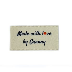 May include: A beige fabric label with the text "Made with love by Granny" in navy blue lettering and a small red heart.