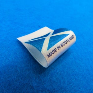 Puede incluir: Una etiqueta blanca con el texto "MADE IN SCOTLAND" y la bandera escocesa, una X blanca sobre un fondo azul. La etiqueta es curva y está colocada sobre un fondo azul.