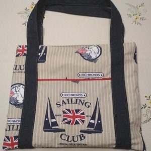 Può includere: Una borsa tote in tela beige con manici in denim blu. La borsa ha un motivo a righe e presenta una bandiera britannica rossa, bianca e blu, barche a vela e il testo "Richmond Sailing Club, Londra, Gran Bretagna, Dal 1923".
