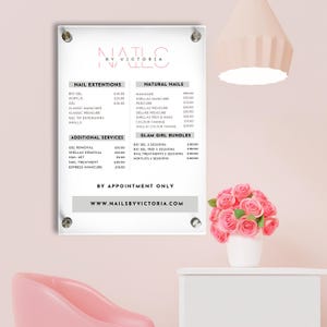 Puede incluir: Una lista de precios enmarcada para servicios de uñas, titulada "Nails by Victoria", se muestra en una pared rosa. La lista incluye servicios como extensiones de uñas, uñas naturales y servicios adicionales.